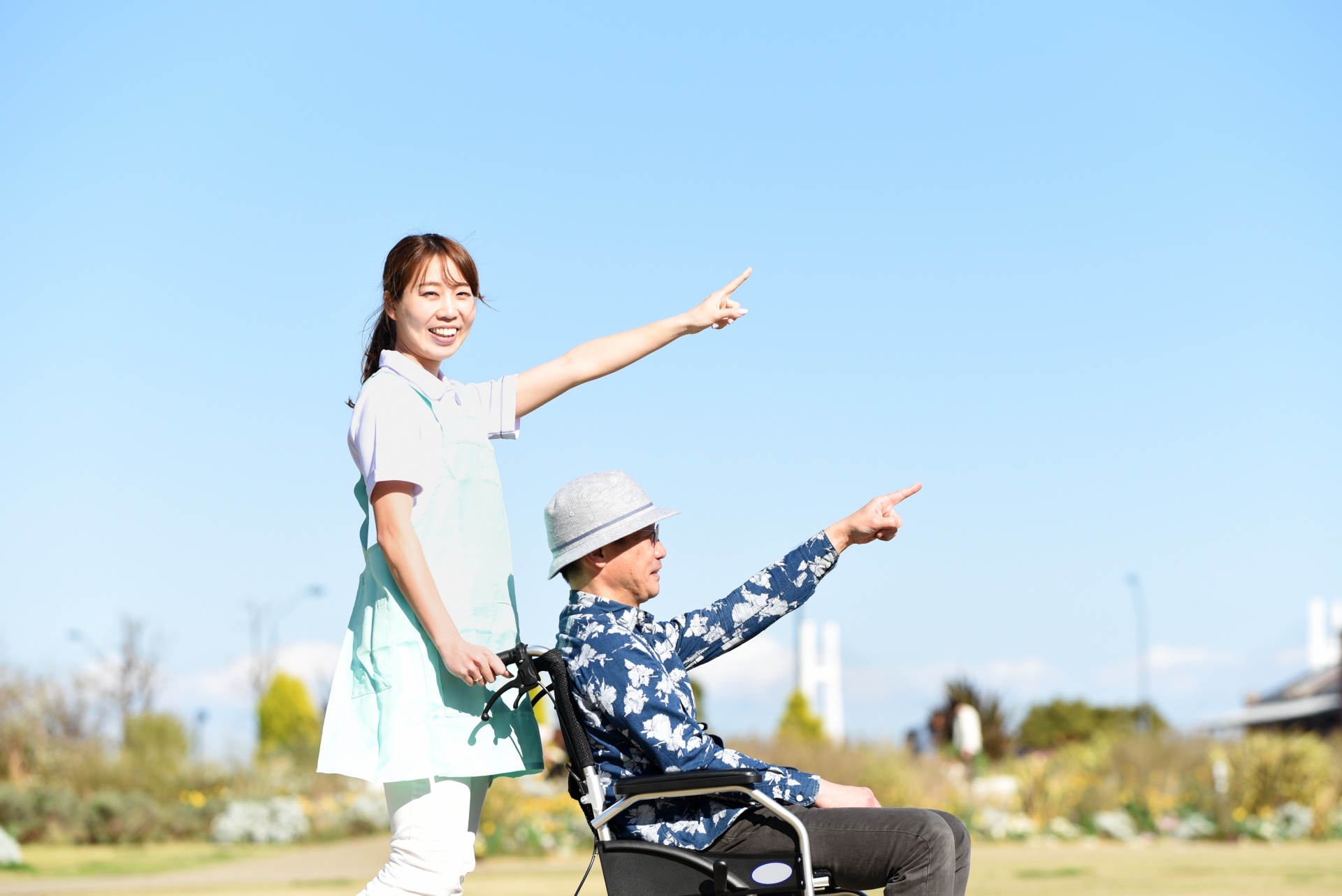 晴れた日に、車椅子に乗った高齢男性と散歩する介護士の女性が、笑顔で前方を指差している様子