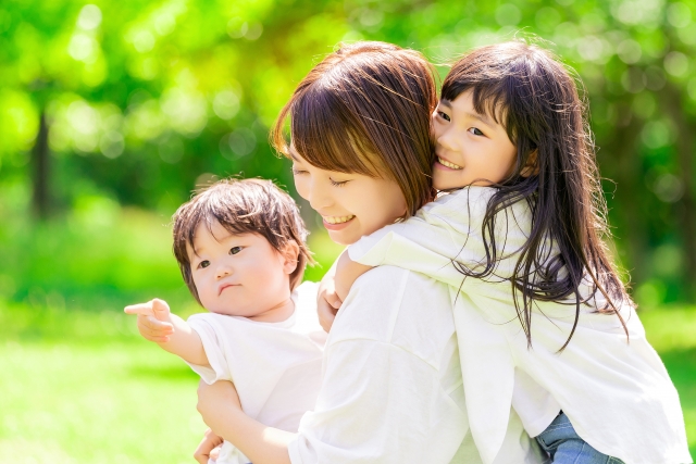 緑豊かな公園で、母親が赤ちゃんを抱っこし、女の子をおんぶしている、笑顔の親子3人