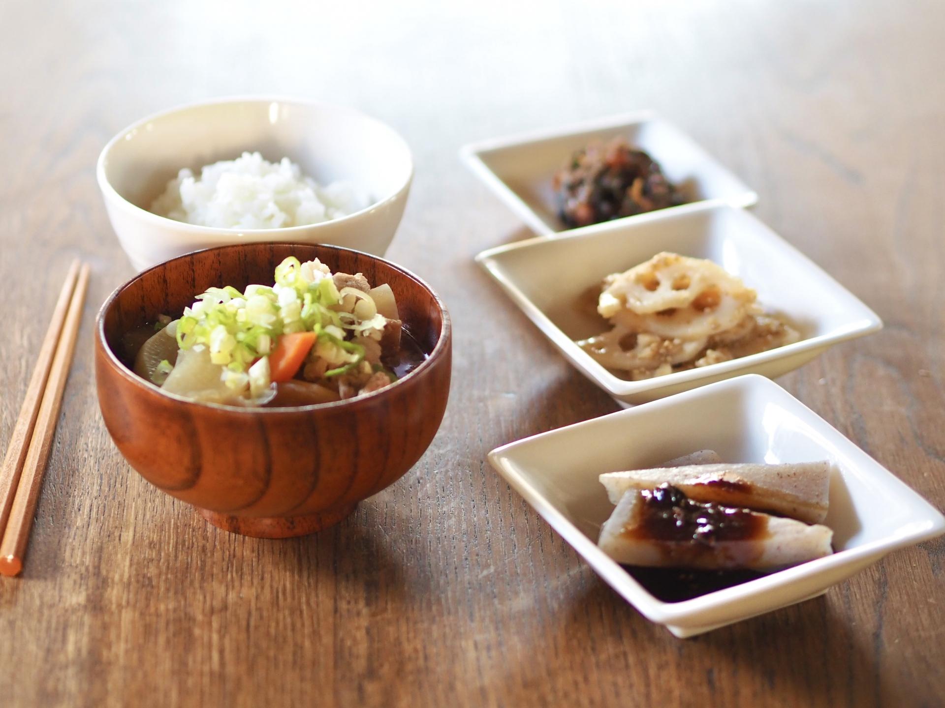 ご飯、豚汁、れんこんのきんぴら、こんにゃくの煮物、佃煮のような小鉢が並ぶ、木のテーブルに置かれた日本の食卓のクローズアップ