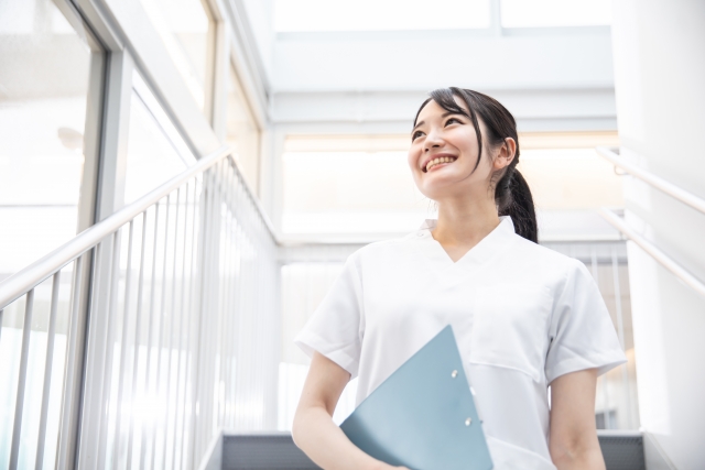 明るい階段でクリップボードを手に、笑顔で見上げる白衣姿の女性看護師