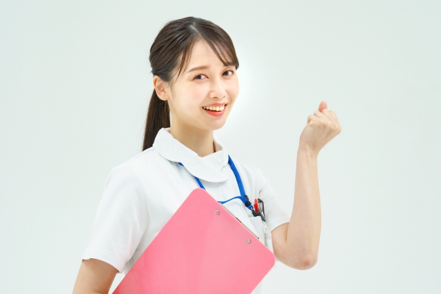 ピンクのクリップボードを抱え、笑顔でガッツポーズをする白衣姿の女性看護師