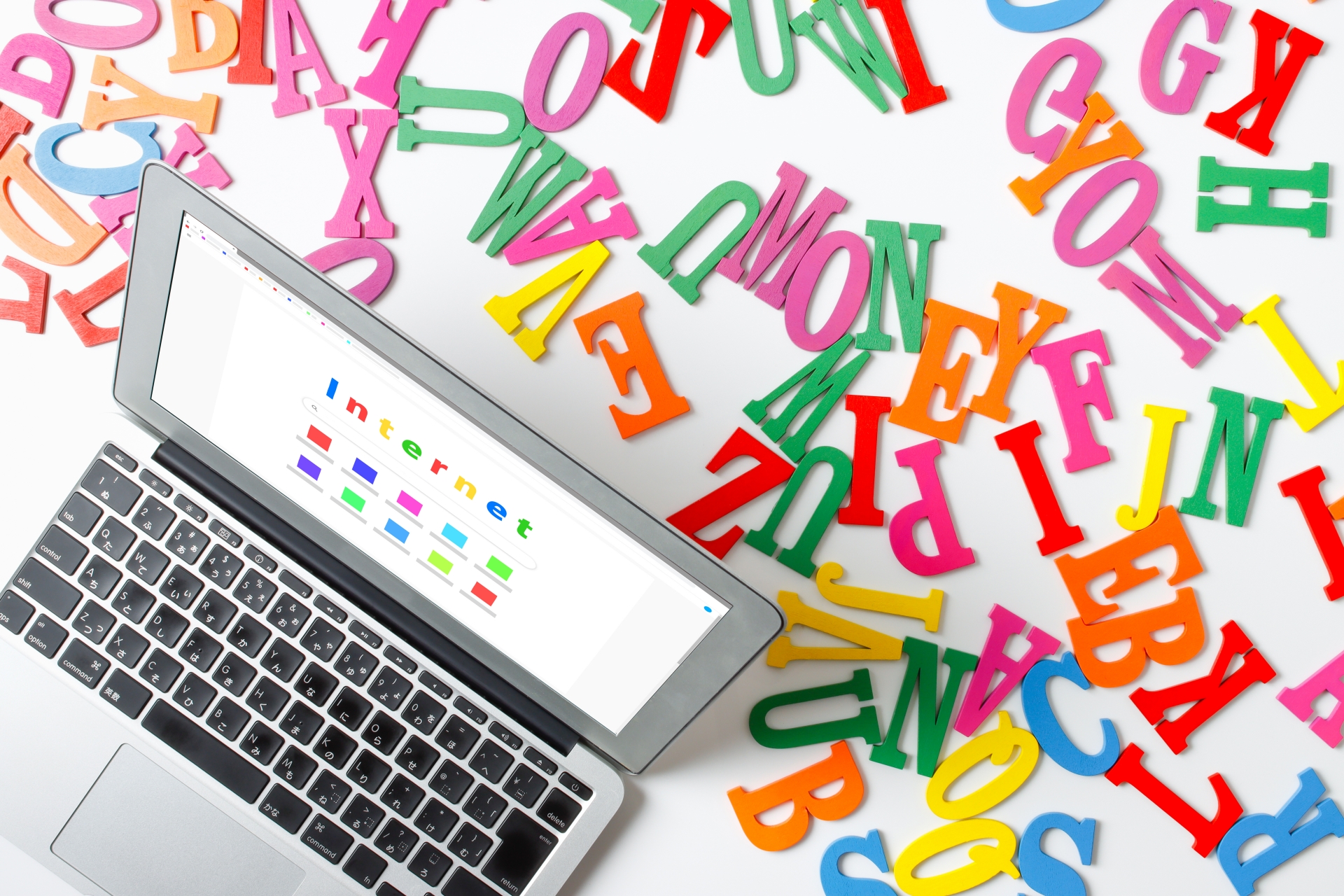 白い背景の上に、検索画面を表示したシルバーのノートパソコンと、散らばった色とりどりのアルファベットの文字ブロックが配置されている俯瞰写真。画面の検索窓には「internet」とカラフルな文字で入力されている。