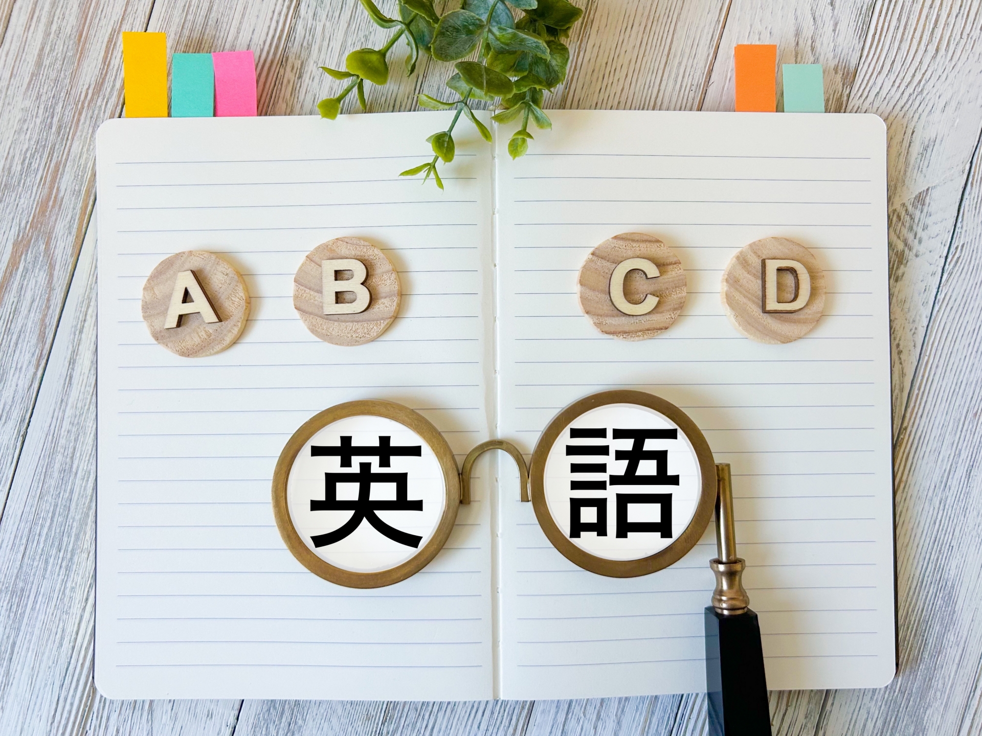 木目調のデスクに開かれたノートの上に、A・B・C・Dと書かれた4つの丸い木製チップが並んでいる。手前には丸いレンズの眼鏡が置かれ、レンズ越しに「英語」という大きな文字が見える俯瞰写真。ノートの上部にはカラフルな付箋と観葉植物が添えられている。