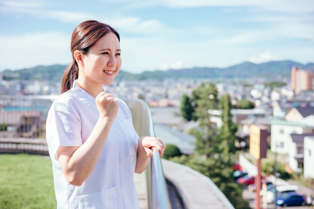 街並みと山を望む屋上で、手すりに手を添え、笑顔でガッツポーズを作る白衣姿の女性看護師