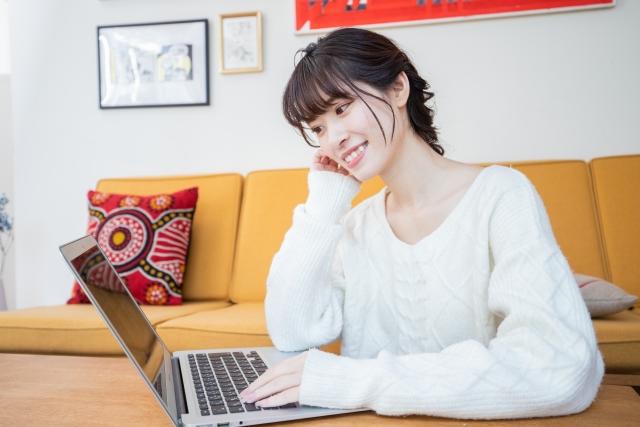 自宅のリビングで、白いニットを着た笑顔の女性が、ローテーブルに置かれたノートパソコンを操作している様子。背景には黄色いソファとインテリアが写っている。