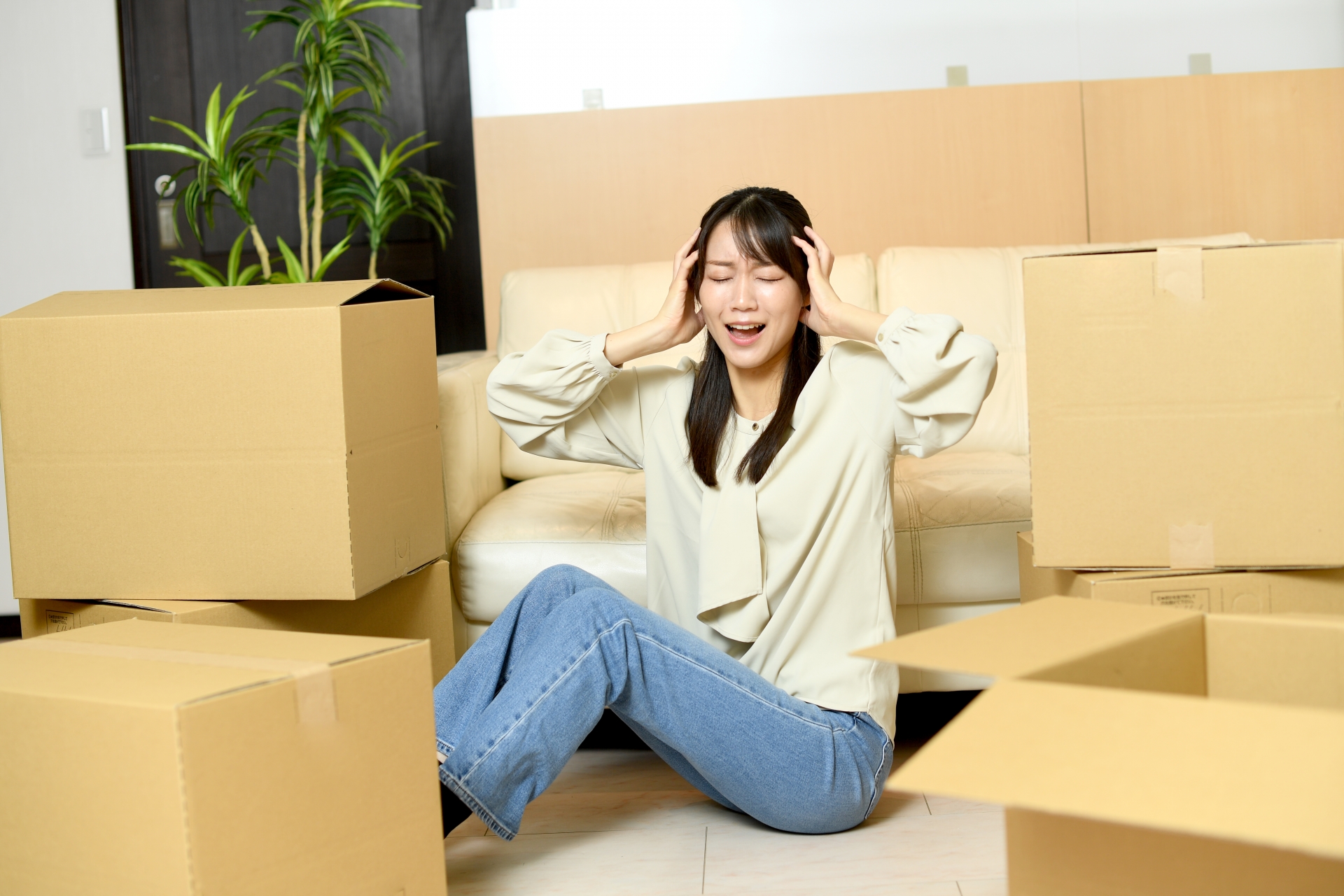 リビングの床に座り込み、両手で頭を抱えて絶望的な表情を浮かべる女性。周囲には荷物が入った大量の段ボール箱が積み上げられており、引っ越し準備の苦労やストレスを感じさせるシーン。背景には白いソファと観葉植物が見える。