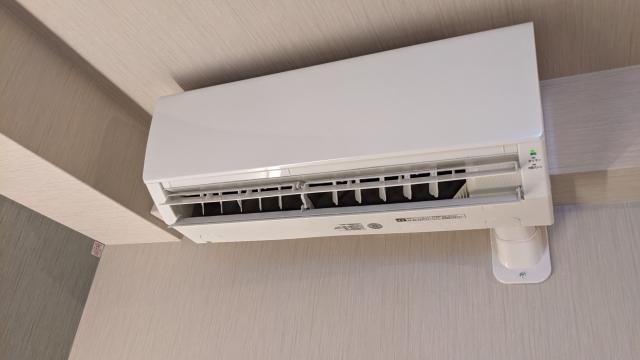 木目調の壁と天井に設置された、清潔感のある白い壁掛け型エアコン。フラップ（ルーバー）が開いており、右下の運転ランプ（緑色）が点灯している状態。