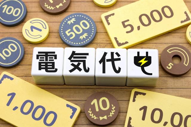 「電気代」の文字とコンセント・雷マークが描かれた白いブロック。その周囲におもちゃの硬貨や紙幣が散りばめられている様子