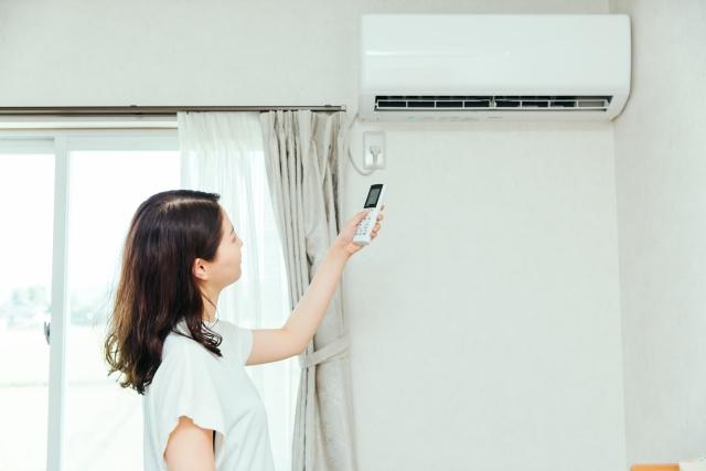 明るい部屋の窓辺で、壁掛けエアコンに向けて白いリモコンをかざして操作している女性の横顔