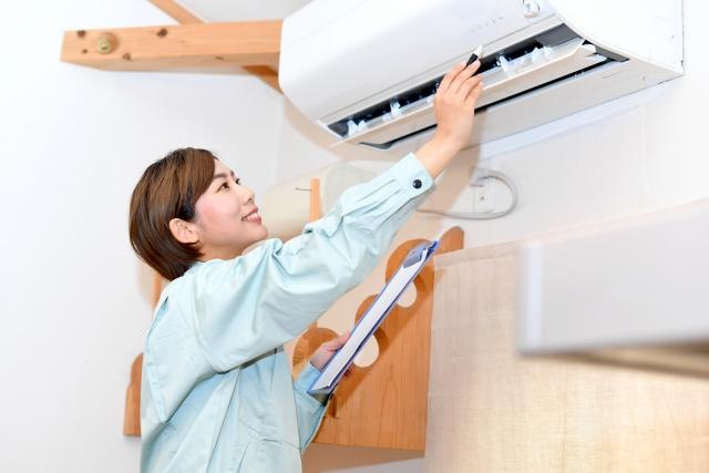 薄いグリーンの作業服を着たショートヘアの女性スタッフが、笑顔でバインダー（クリップボード）を抱えながら、壁掛け型エアコンの吹き出し口（ルーバー）を指先でチェックしている点検作業のイメージ画像。