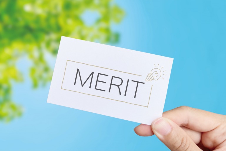 「MERIT」と書かれたカードを持つ手と、背景に緑の葉と青空