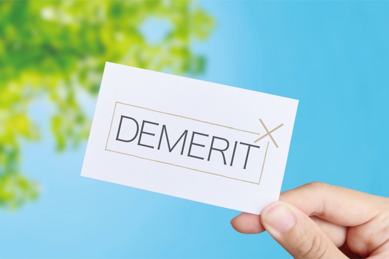 「DEMERIT」と書かれたカードを持つ手と、背景に緑の葉と青空