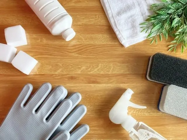 木目調のテーブルの上に、グレーのゴム手袋、スプレーボトル、スポンジ、白いボトル、メラミンスポンジ、白いタオルなどが円を描くように並べられている俯瞰写真。