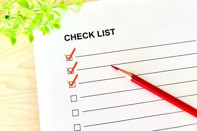 木目調のテーブルに置かれた「CHECK LIST」という紙。上から3つのチェックボックスにオレンジ色のチェックマークが入っており、その横に赤い鉛筆が添えられている。左上には緑の葉が見える。