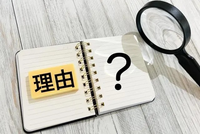 白い木目調のテーブルに置かれたリングノート。左ページには「理由」と書かれた黄色い付箋があり、右ページには大きな「？」マーク。ノートの横には虫眼鏡が置かれている。