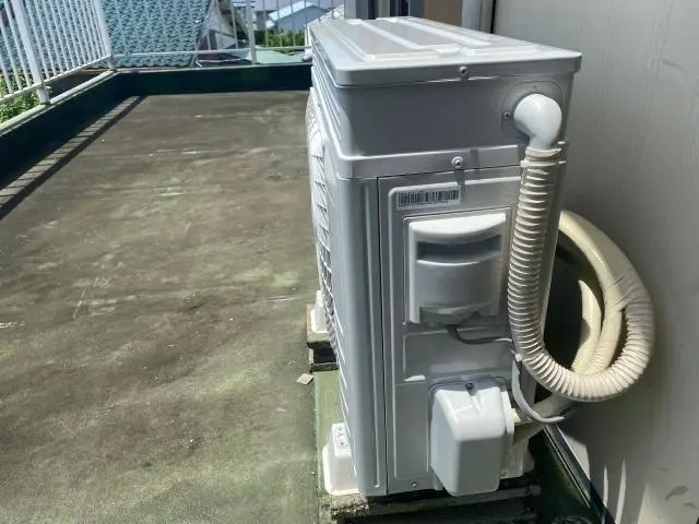 ベランダに設置された白いエアコン室外機の、配管が接続されている右側の側面を写したクローズアップ写真。室外機から太い蛇腹状のホースが伸び、壁に沿ってまとめられている。