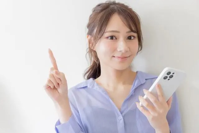左手に白いスマートフォンを持ち、右手で人差し指を立ててポイントを指し示す笑顔の女性。紫色のシャツを着用し、シンプルな白い背景の前に立っている。