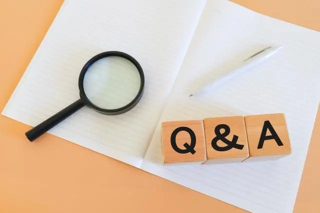 開いた白いノートの上に、「Q」「&」「A」と書かれた3つの木製ブロックが並んでいる。ノートの左側には黒い虫眼鏡、右側には白いペンが置かれている。