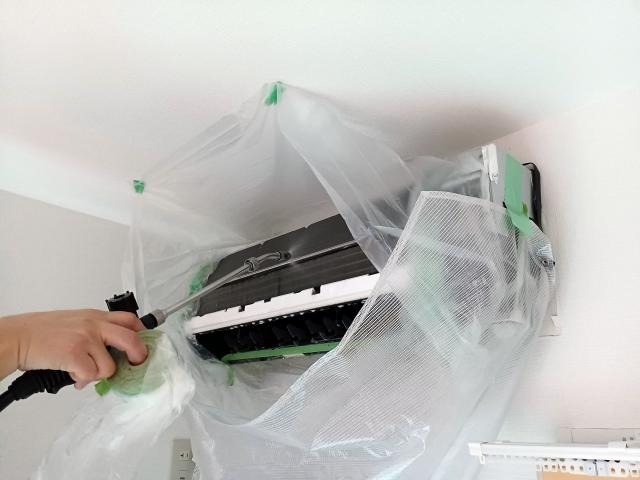 ビニールシートで養生された壁掛けエアコンの内部に、高圧洗浄機のノズルから洗浄液を噴射して清掃している様子。