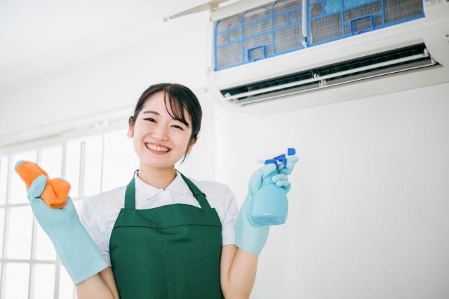 掃除道具のスプレーと布を持ち、壁掛けエアコンの前で満面の笑みを見せる作業服姿の女性スタッフ。