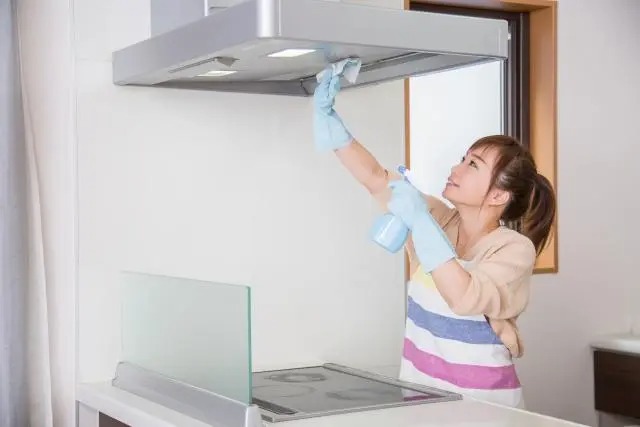 明るいキッチンで、水色のゴム手袋をはめた女性がスプレーと布を使ってレンジフードの内側を拭き掃除している様子。